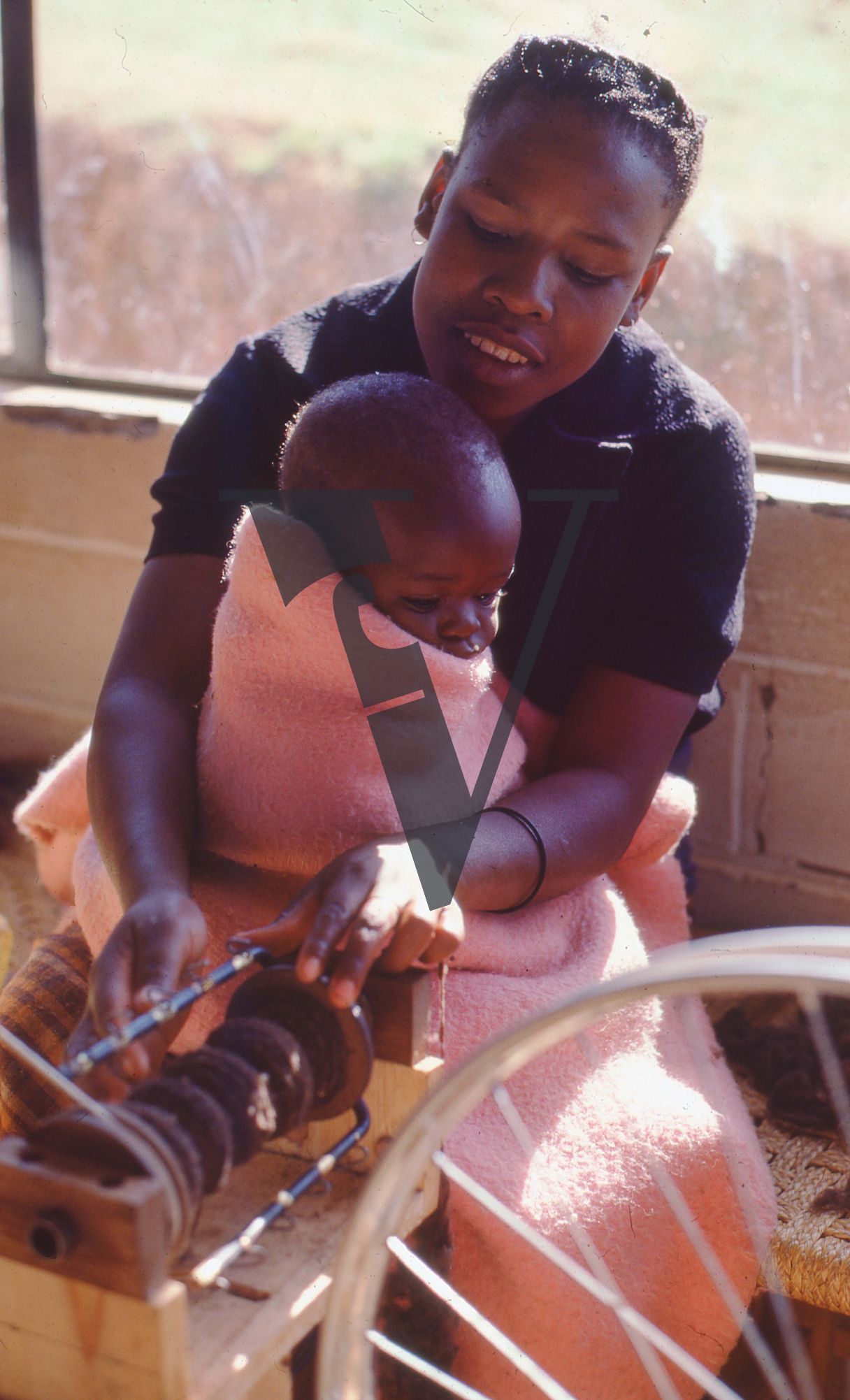 Lesotho, Handspun mohair farming community, spinning wheel, woman at work, holds child in blanket.