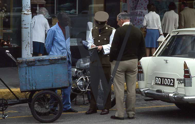 Rhodesia, Salisbury, traffic policeman issuing a roadside ticket, two men, parked cars, street scene.