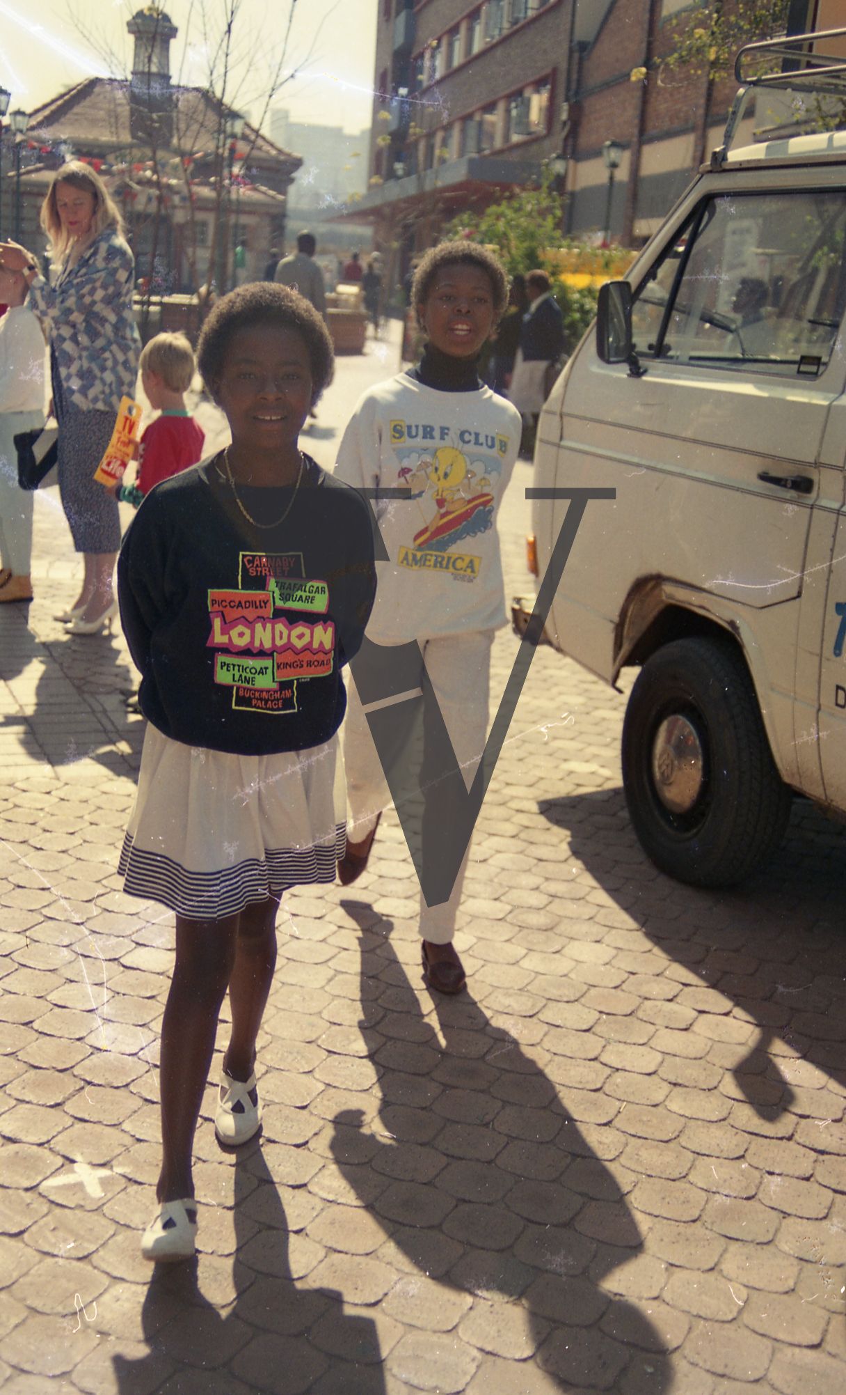 South Africa, Soweto, street scene, two Black girls in foreground, smiling, figurative, white family in background, van, people.