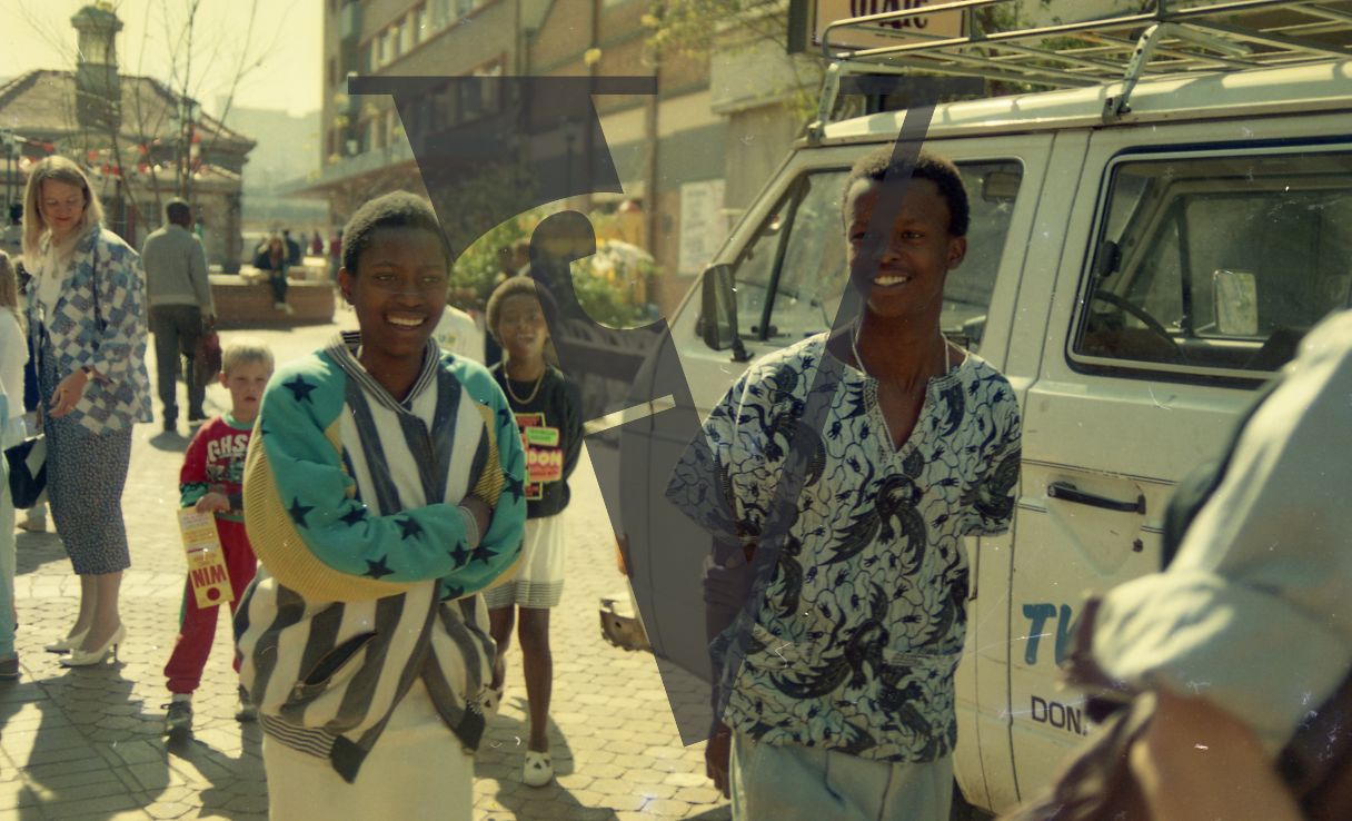 South Africa, Soweto, street scene, two young Black adults, smiling, mid-shot, white family in background, van, people.