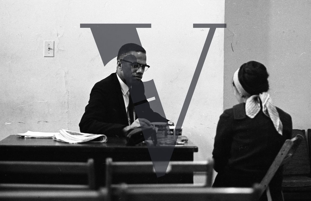 Malcolm X, speaking to woman, at desk, close.